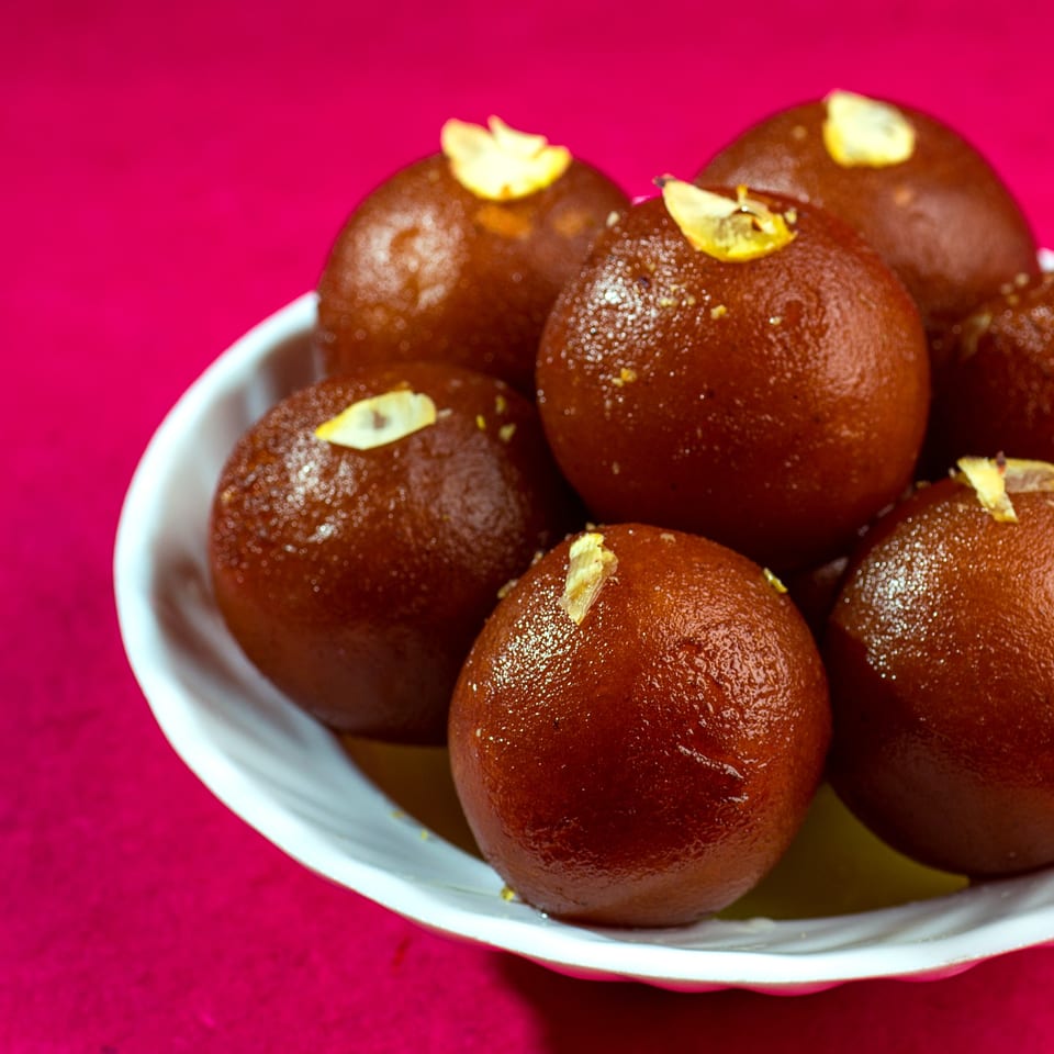 A white bowl filled with glistening brown gulab jamun, garnished with slivered almonds, on a pink background.