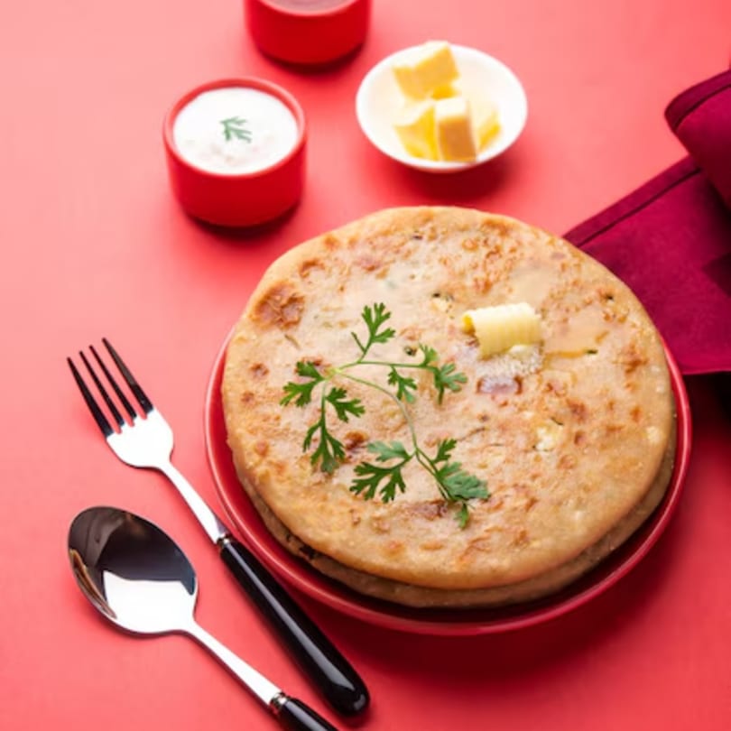 Indian paratha with butter and cilantro, served with yogurt and chutney on a red background.