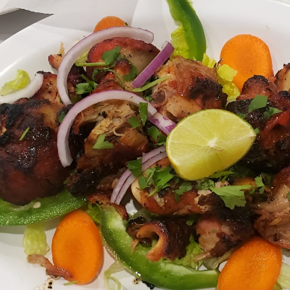 Indian grilled chicken with red onions, cilantro, lime, green peppers, and carrots on a white plate.